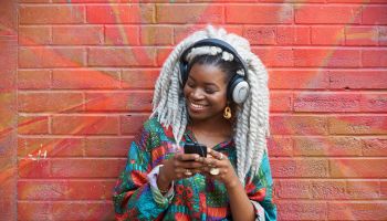 Black woman leaning on brick wall listening to cell phone with headphones