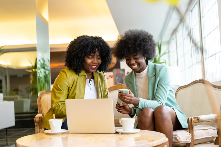 Black ethnic girls in a hotel cafeteria. African women with curly and afro hair, looking at the mobile with some coffee