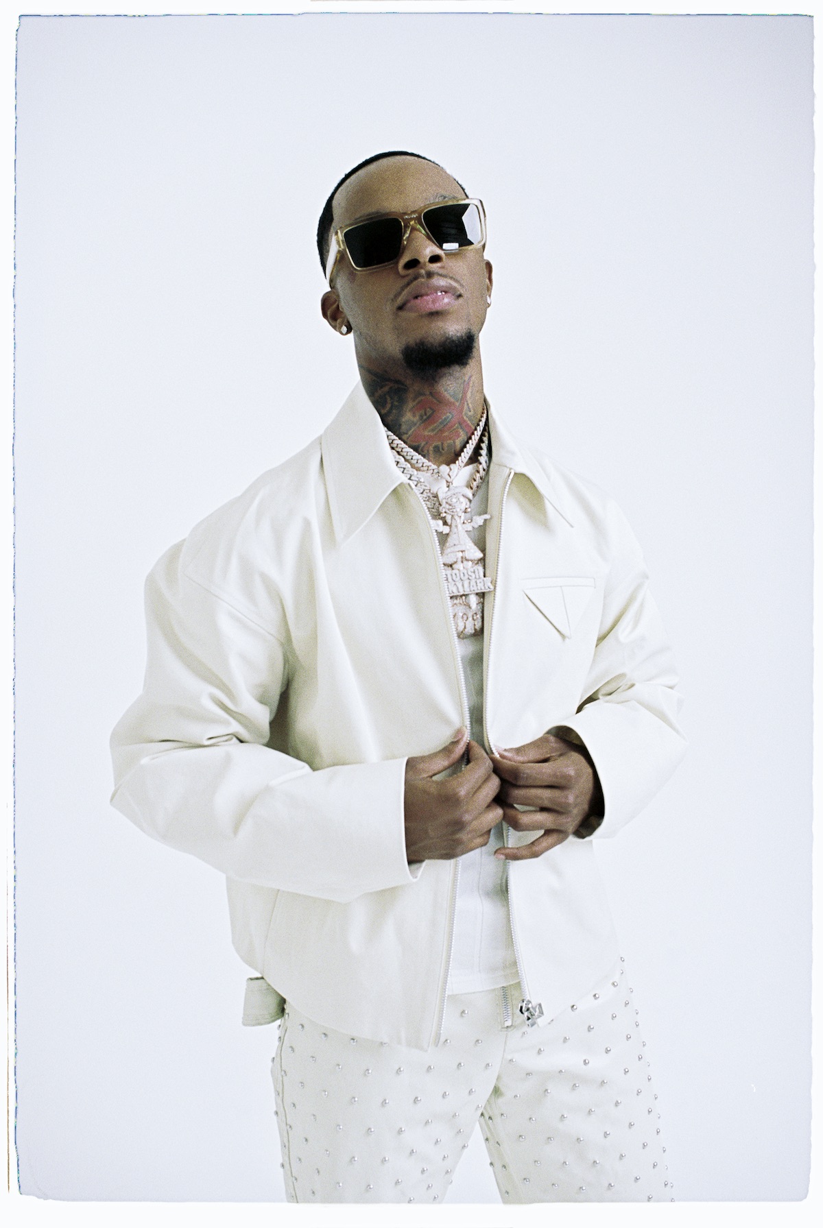 A man wearing sunglasses and a white jacket stands against a plain background.