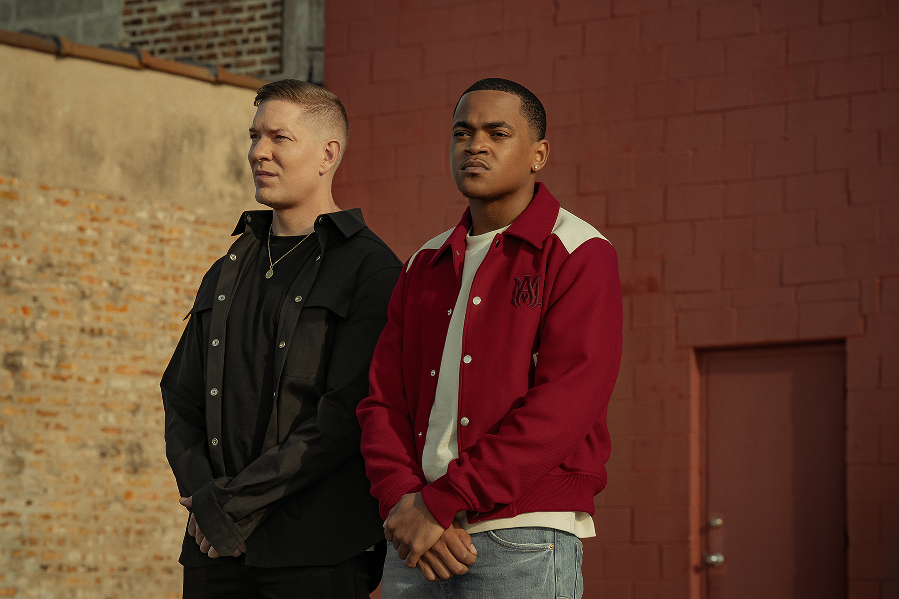 Two young men standing in front of a red brick wall, one wearing a black jacket and the other a red jacket.