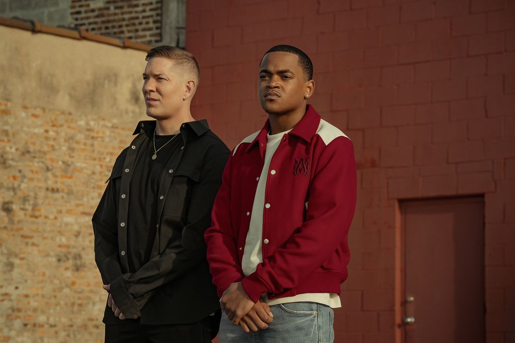 Two young men standing in front of a red brick wall, one wearing a black jacket and the other a red jacket.