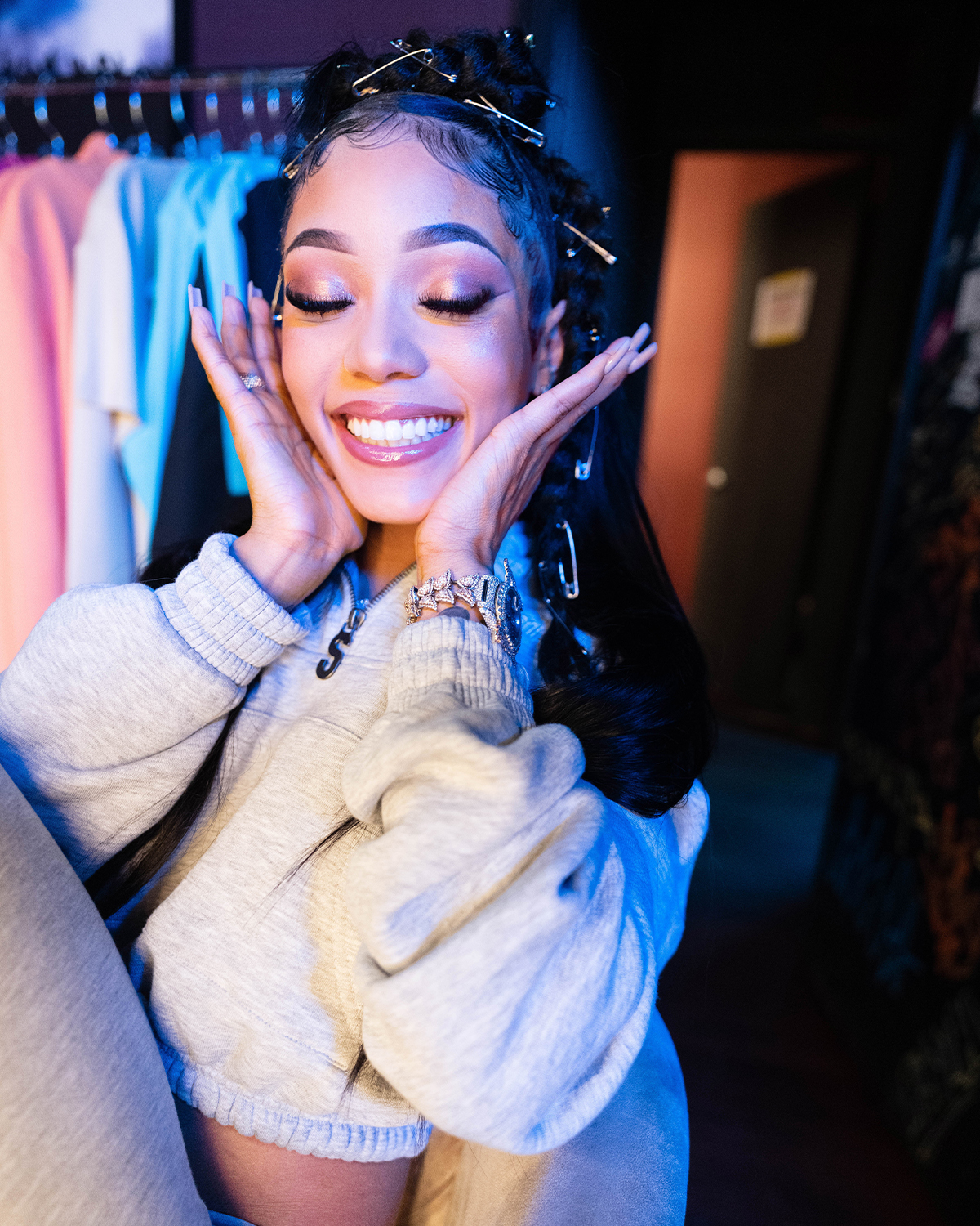 A smiling woman with elaborate hairstyle and makeup, wearing a cream-colored sweater, posing in front of a colorful clothing display.
