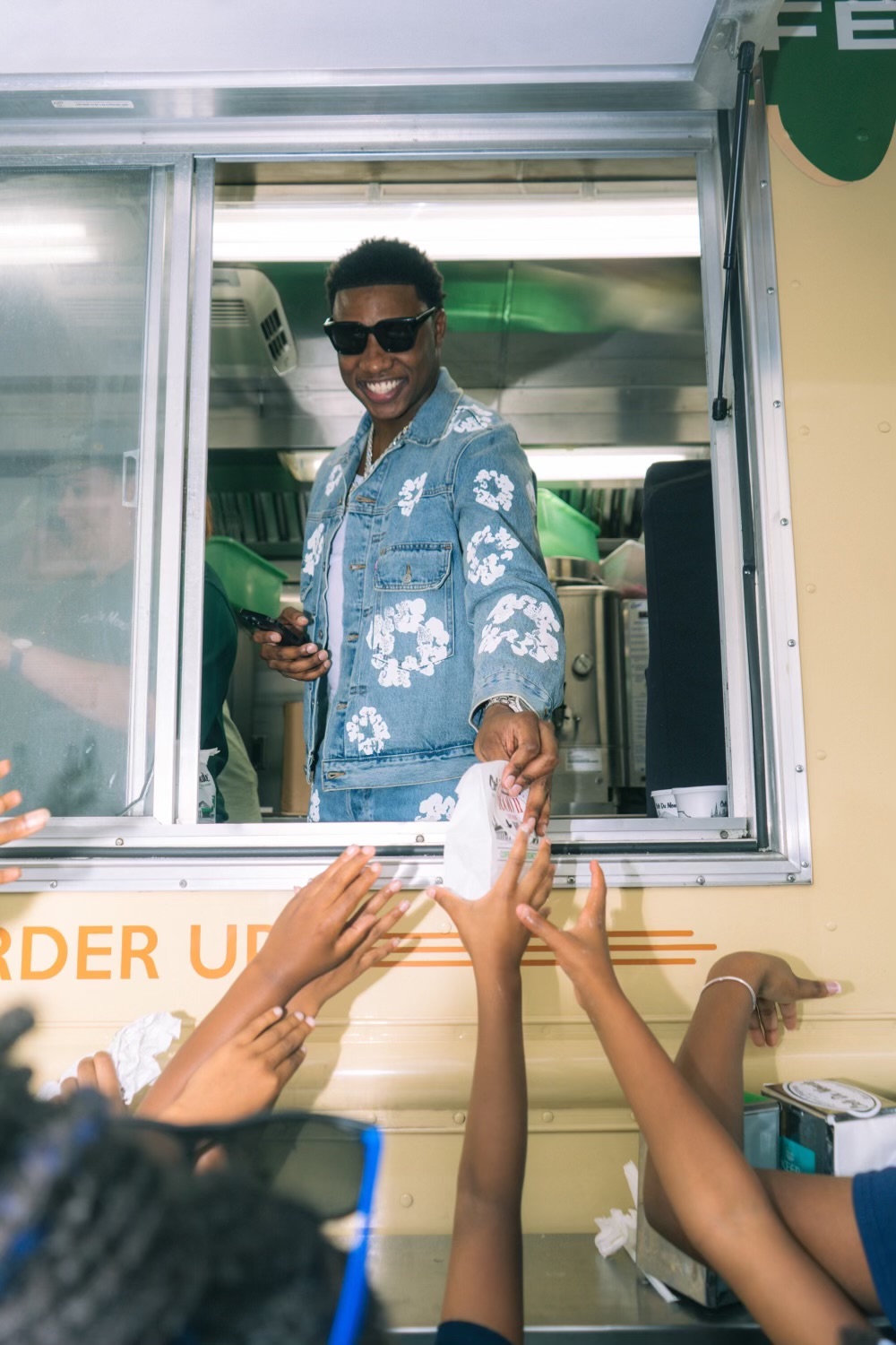 A smiling person in a denim jacket with floral designs stands in a food truck window, handing something to outstretched hands.