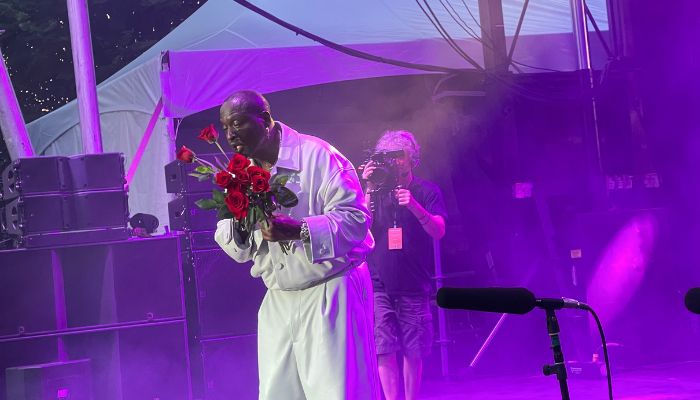 A person in a white robe holding a bouquet of red roses on a stage with purple lighting and smoke effects.