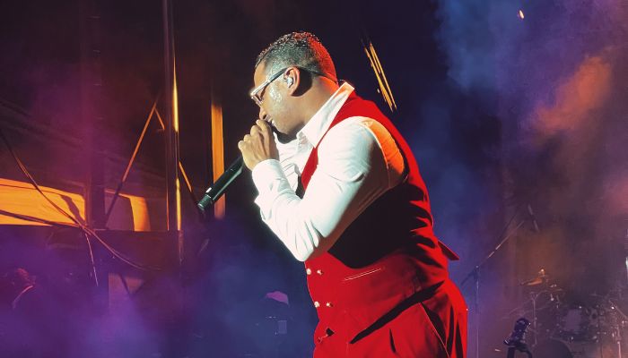 A performer in a red suit singing into a microphone on stage with smoke and colorful lighting.