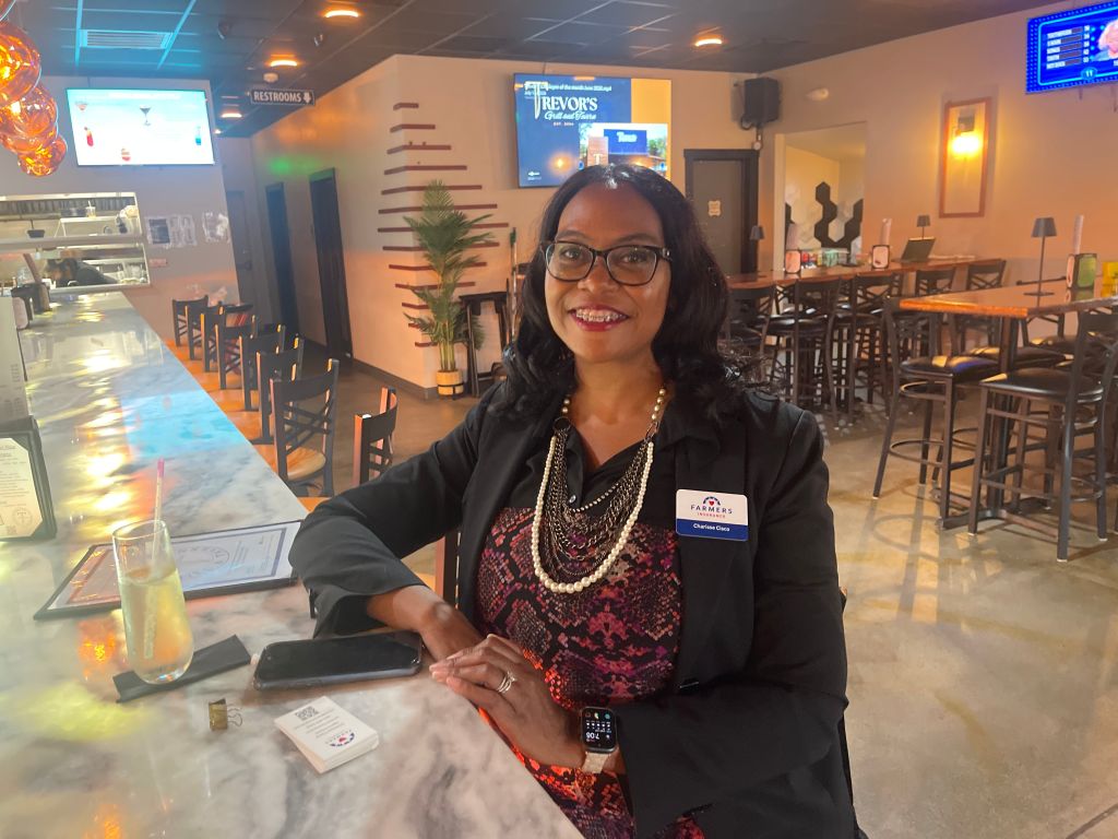 A smiling woman wearing glasses and a necklace stands behind a counter in a restaurant or cafe setting, with a Trevor's logo visible on the wall.