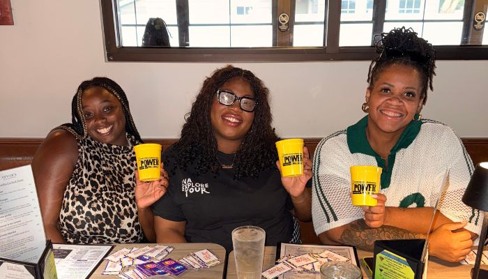 Three smiling women holding yellow mugs with "KWEH" printed on them, sitting at a table with various items.