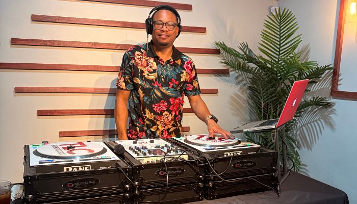 A man wearing headphones and a floral shirt stands behind a DJ mixer, surrounded by plants in a studio setting.