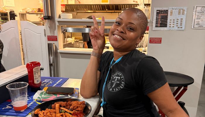 A smiling woman wearing a black t-shirt with a logo, making a peace sign gesture while sitting at a table with food.