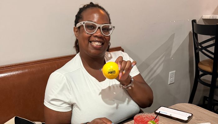 A smiling person wearing glasses and holding a yellow tennis ball.