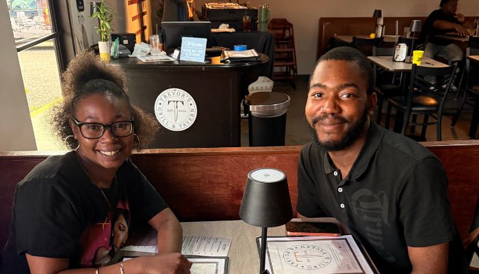 Two people, a man and a woman, sitting at a table in what appears to be a cafe or coffee shop. They are smiling and appear to be engaged in conversation.
