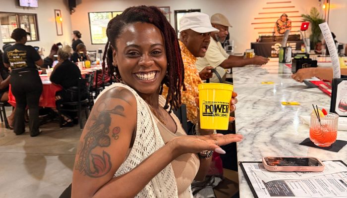 A smiling woman with dreadlocks holding a yellow cup with "KWET" printed on it, seated in a restaurant setting with other people in the background.