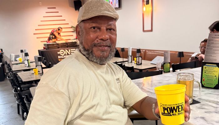 An older man with a gray beard wearing a cap and a white shirt, holding a yellow cup with the word "Power" on it, sitting in what appears to be a restaurant or cafe setting.