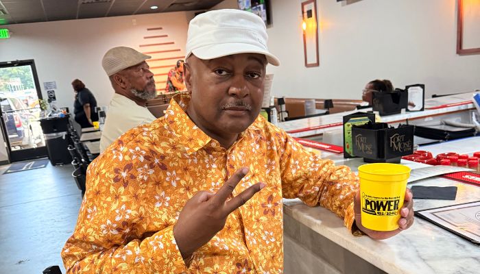 A man wearing a yellow floral shirt and a white hat standing in a room, holding a yellow container labeled "Power".