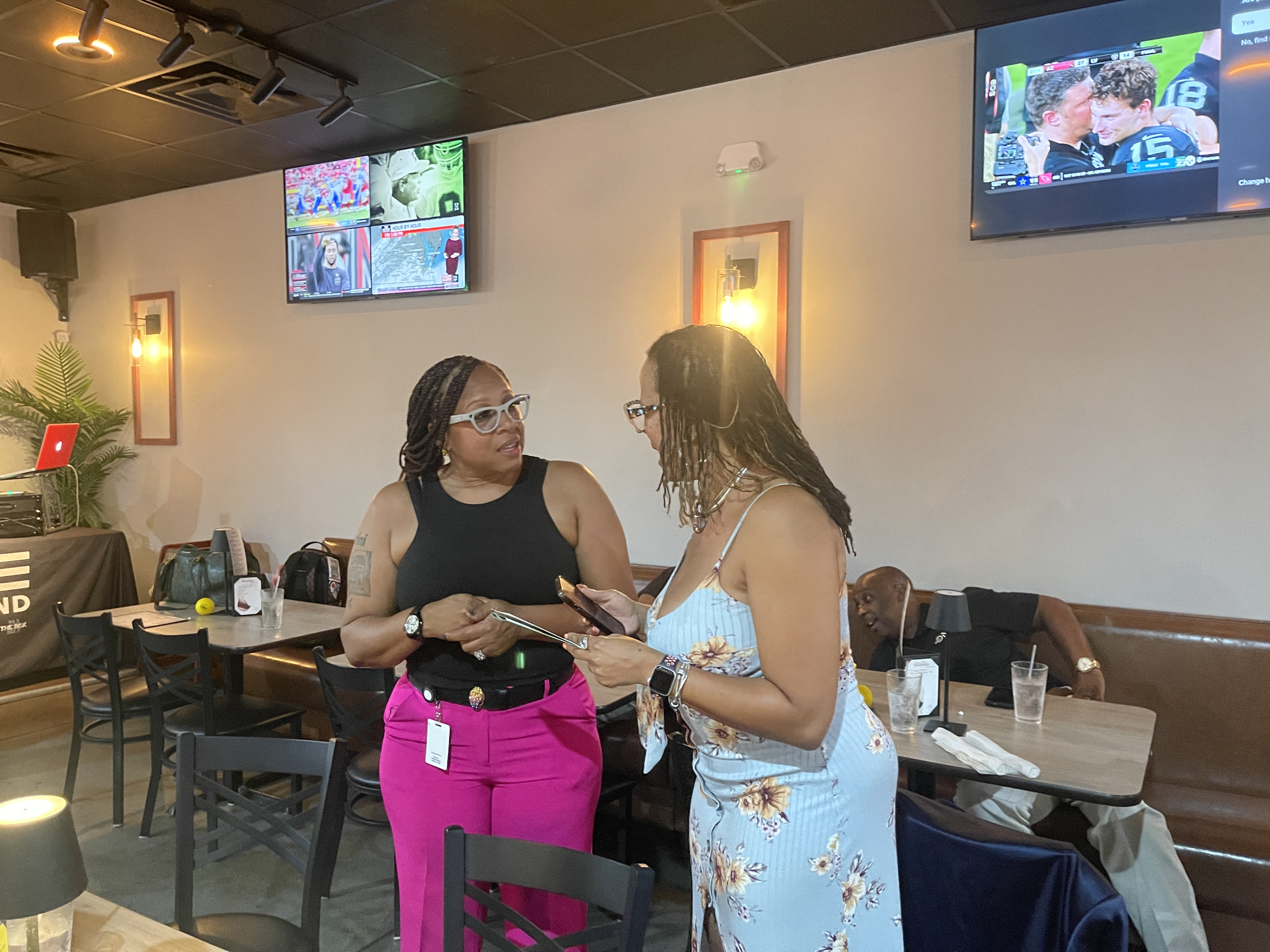 Two women in casual clothing conversing at a table in a restaurant or cafe, with televisions displaying sports programming on the walls behind them.