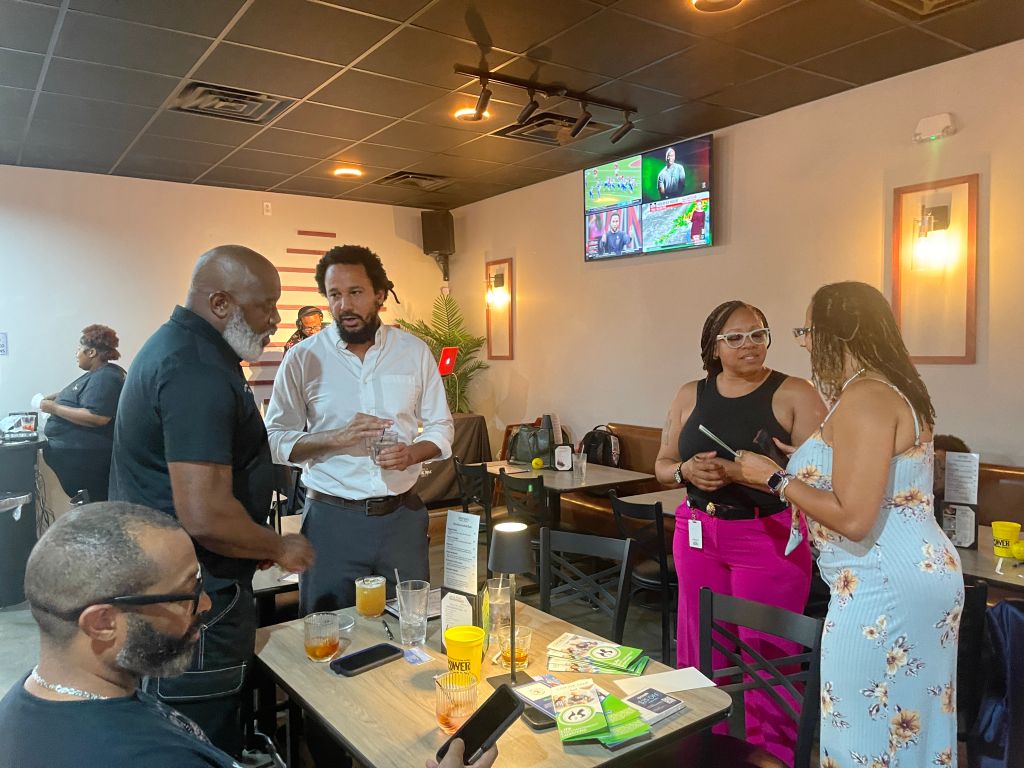 A group of people conversing in a casual dining setting, with a television screen displaying sports programming in the background.