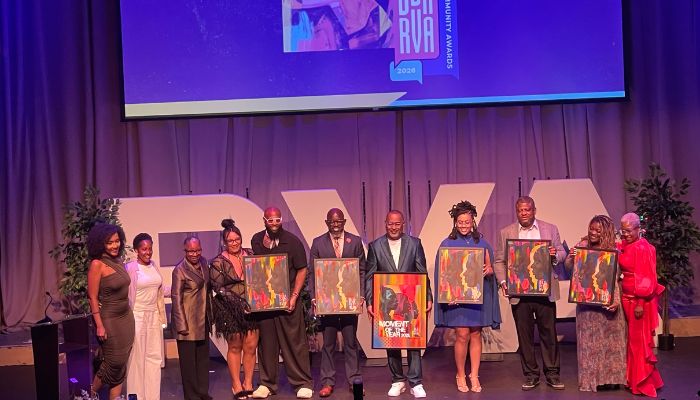 A group of people holding colorful artwork on stage at an event, with a large screen displaying event branding in the background.