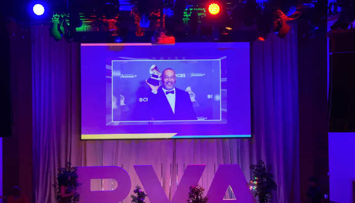 A large screen displays a man in a tuxedo at what appears to be an awards show or event, with colorful lighting and decorations surrounding the stage.