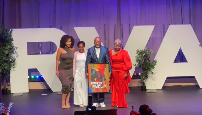 Four people standing on stage, two women in colorful dresses and two men in suits, holding a framed artwork.