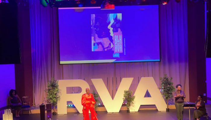 A person in a red outfit standing on a stage with large "RVA" letters and a large screen displaying abstract graphics.