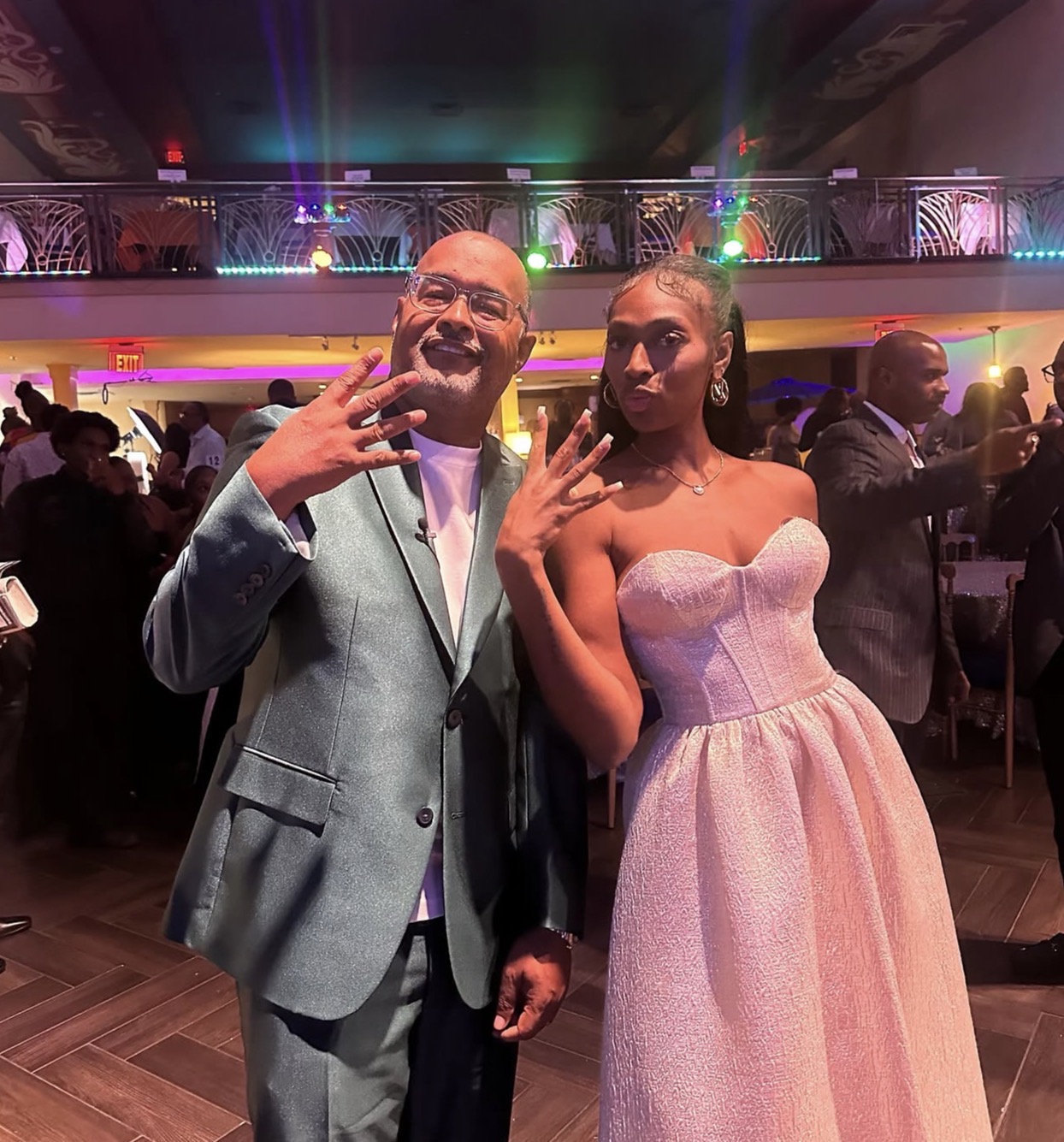 Two people posing at a formal event, with colorful lighting and decorations in the background.