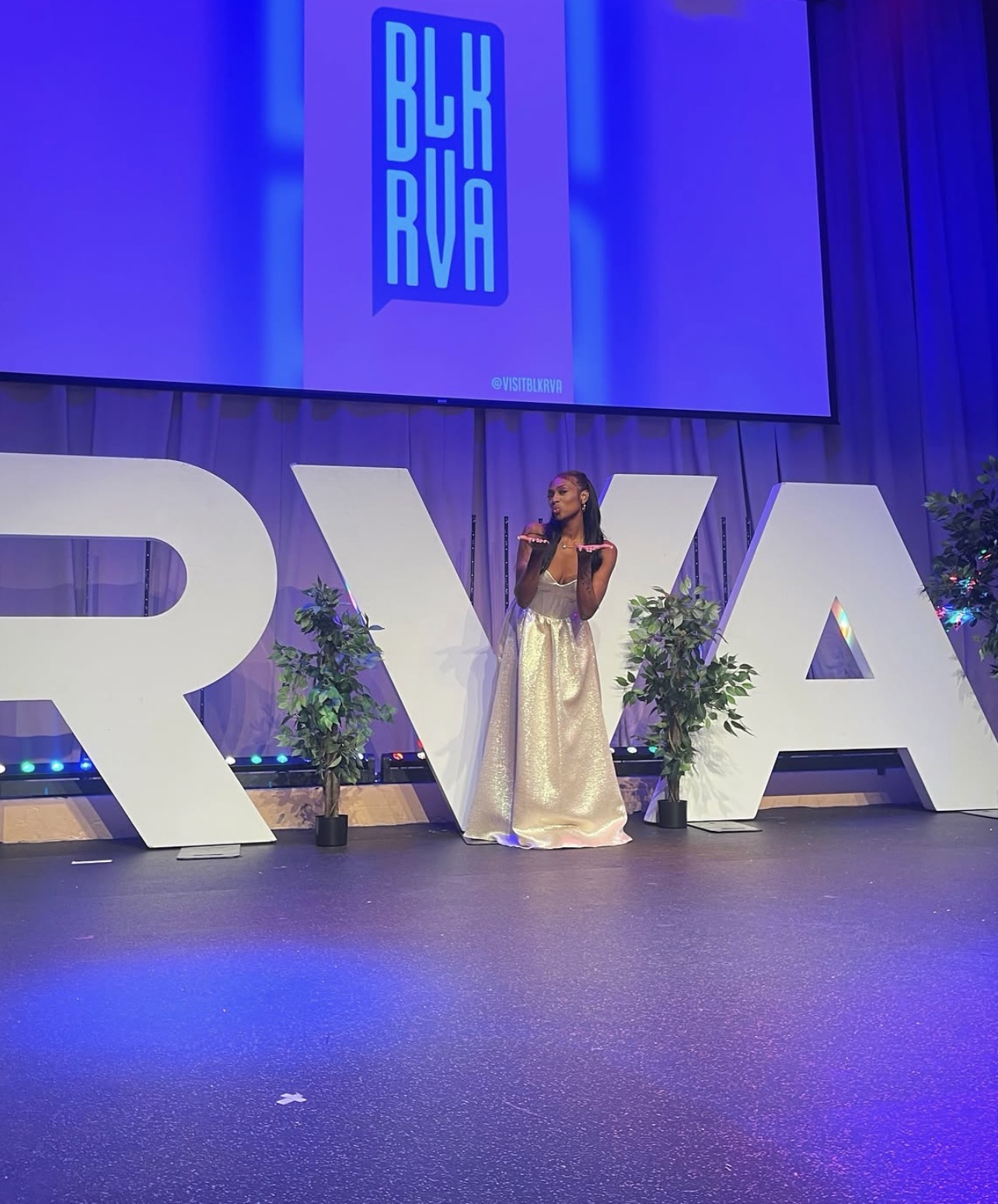 A woman in a gold dress stands on a stage with the text "BURNA" displayed above her.
