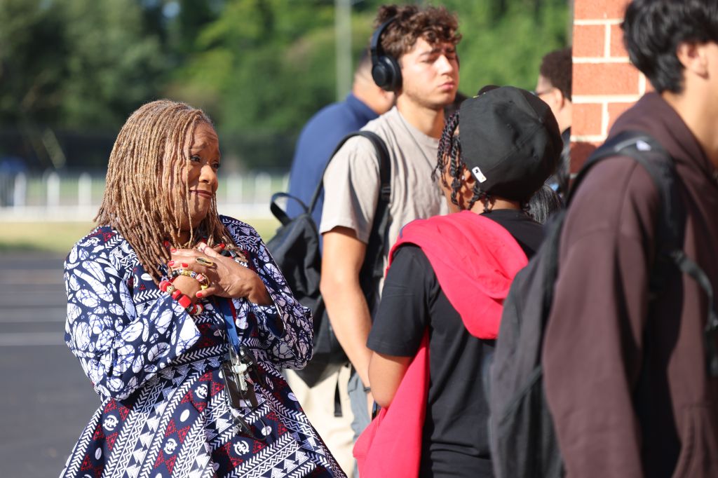 A group of people, including a woman with dreadlocks wearing a patterned dress, standing and conversing outdoors.