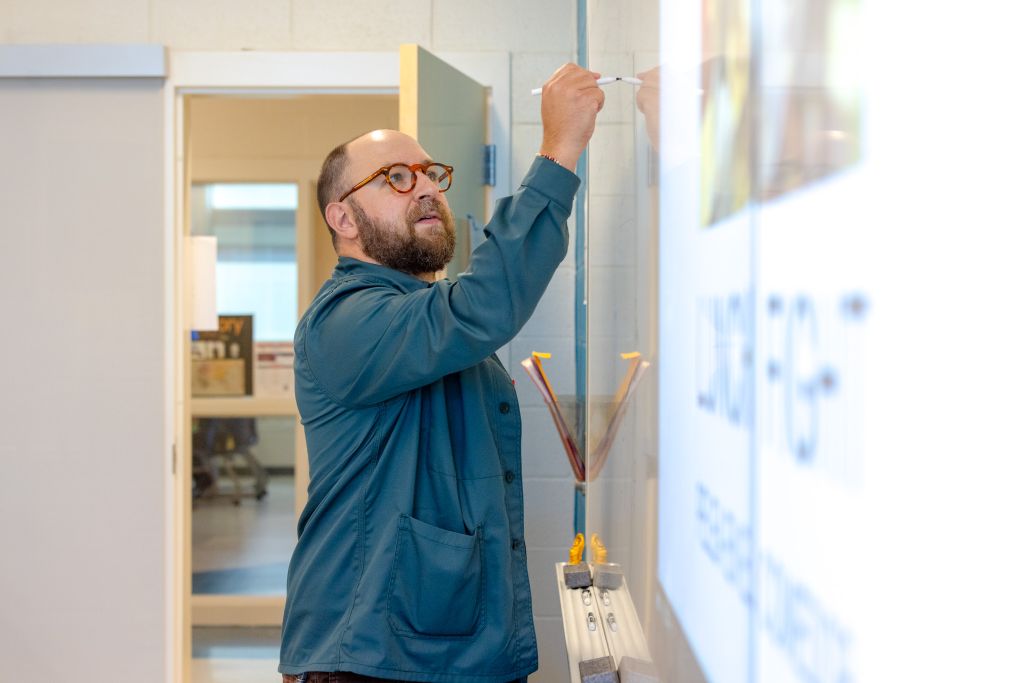 A bearded man in a blue jacket adjusting a whiteboard in an office setting.